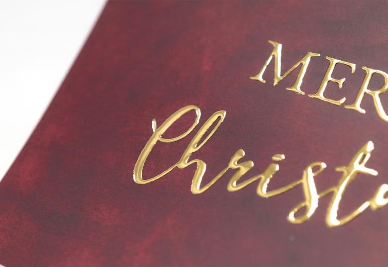 Close-up van een bordeauxrode kerstkaart met de tekst "Merry Christmas" in elegante gouden folieletters, waarbij de feestelijke personalisatie wordt benadrukt.