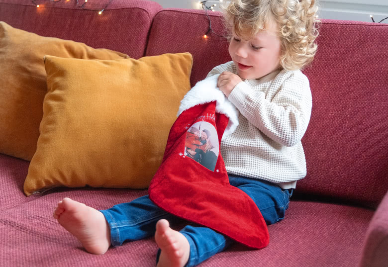 Rød julestok med hvid kant, personliggjort med et foto og "Merry Christmas" tekst, holdt af et barn på en sofa.