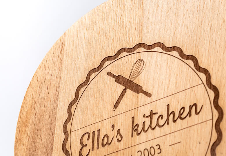 Round wooden cutting board with personalised engraving reading "Ella's kitchen" and a whisk and rolling pin design.