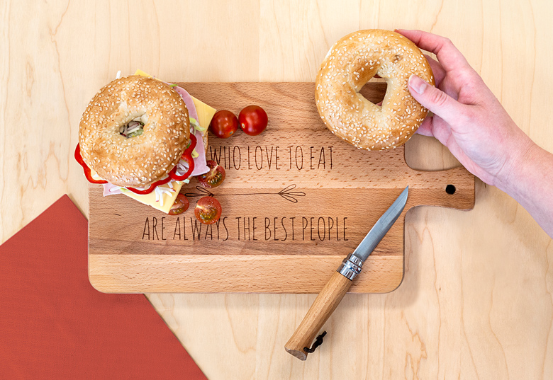 Wooden cutting board with handle, personalised with "WHO LOVE TO EAT ARE ALWAYS THE BEST PEOPLE," shown with bagels, knife, and tomatoes.