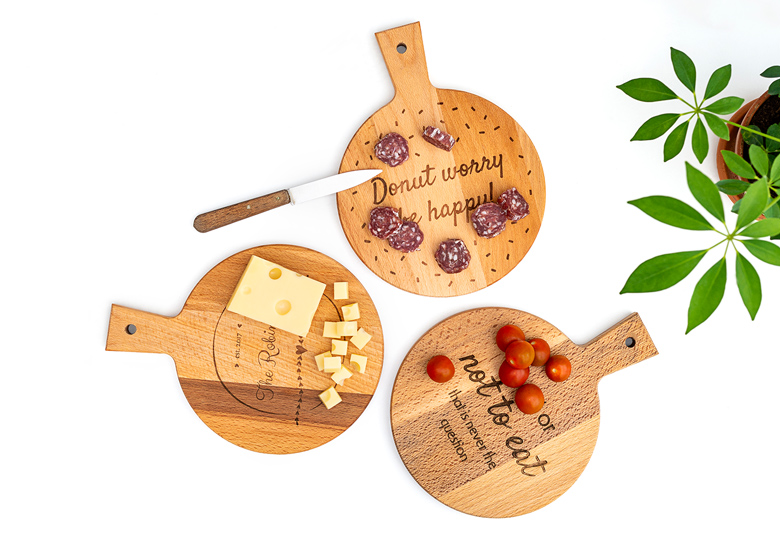 Three round wooden cutting boards with handles, each personalised with different engraved texts, surrounded by food items.