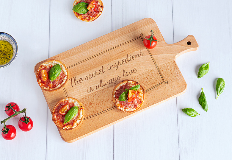 Wooden cutting board with a handle, engraved with "The secret ingredient is always love," decorated with mini pizzas and fresh basil.