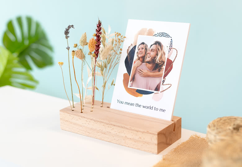 Carte de recharge photo personnalisée avec un design abstrait coloré, displayed in a wooden stand with dried flowers. Text reads: "You mean the world to me."