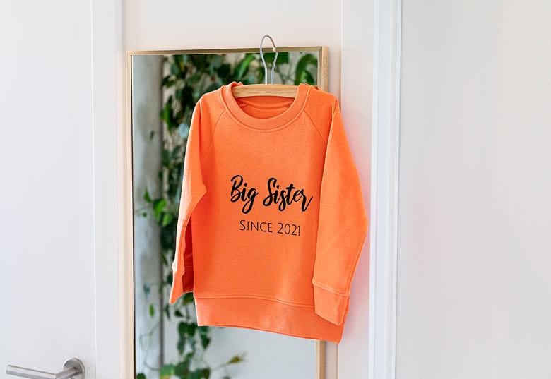 Oranje kindersweater met "Big Sister SINCE 2021" in zwarte letters aan een kapstok.