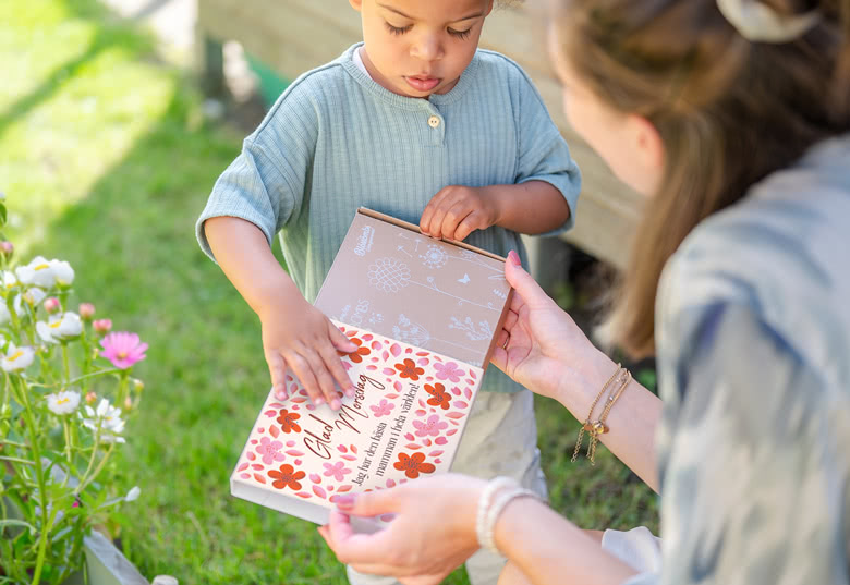 Presentask med vildblomsfröbomber med blommigt design och texten "Glad morsdag" på framsidan, som öppnas av en mamma och ett barn.